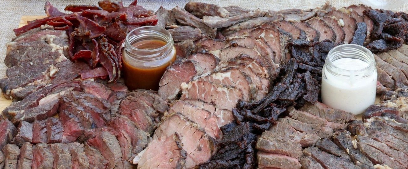 A board of sliced steaks, pastrami and jerky with a jar of sauce at each side