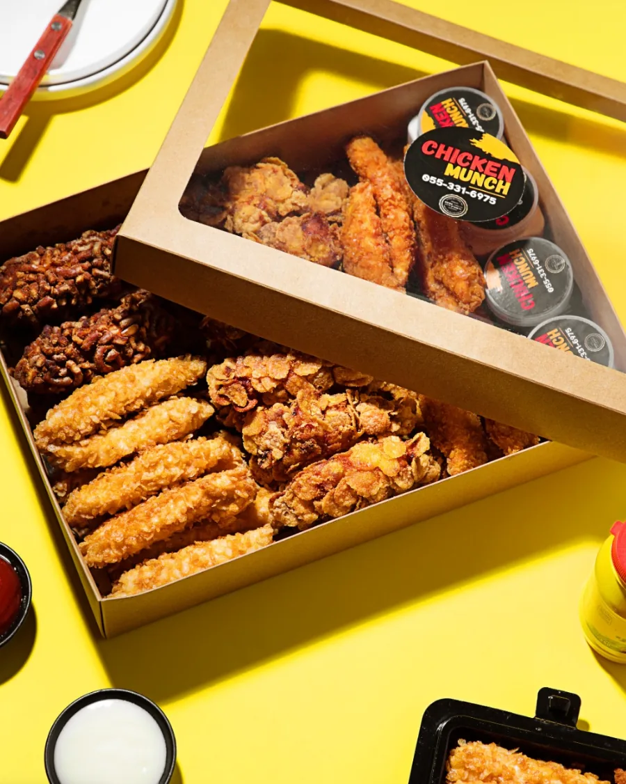 An open Chicken Munch box of pretzel, rice krispie and cornflake chicken, with his labelled sauce pots in the lid, on yellow