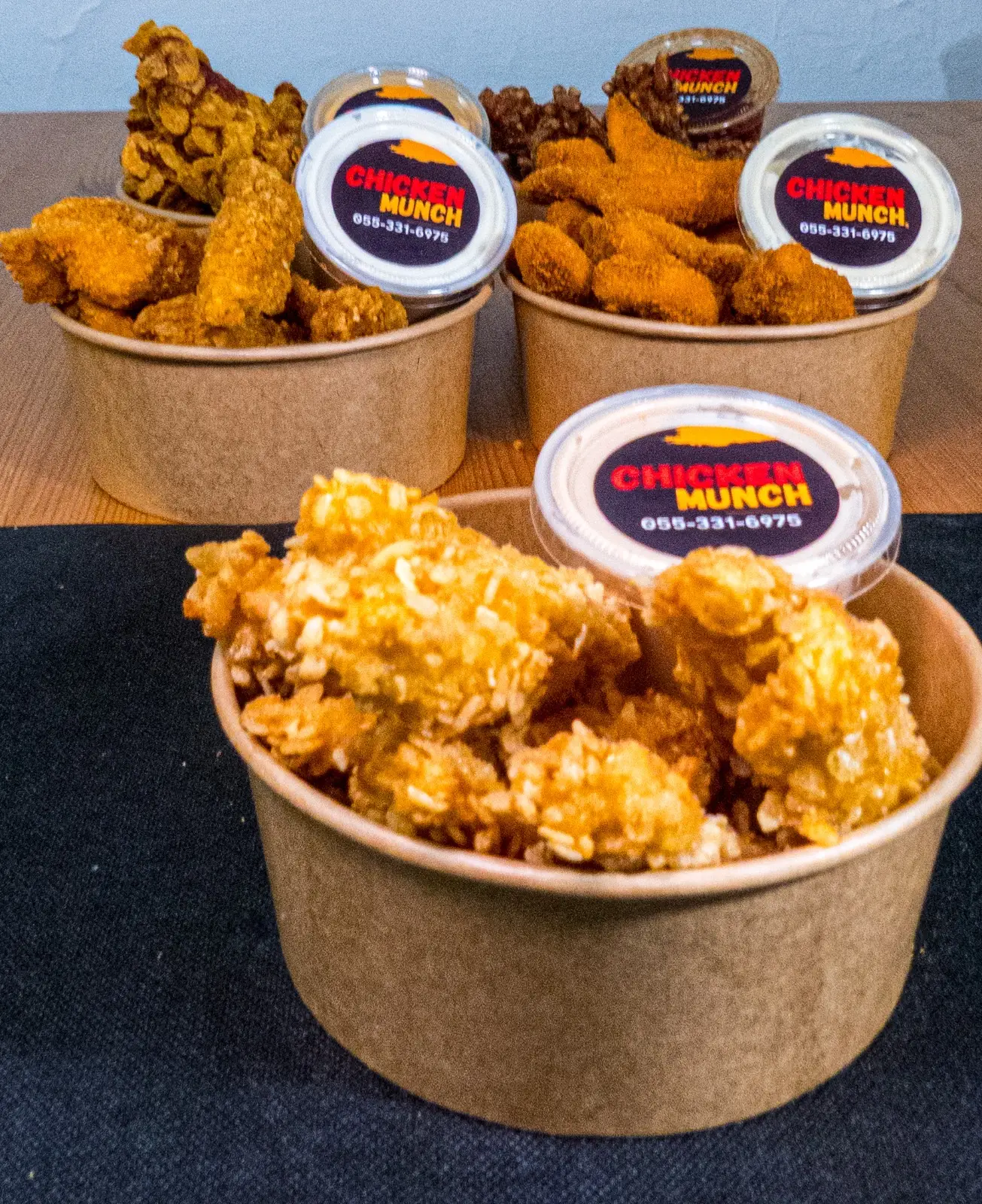 Four kraft bowls of Chicken Munch chicken with sauce pots, on a wooden table