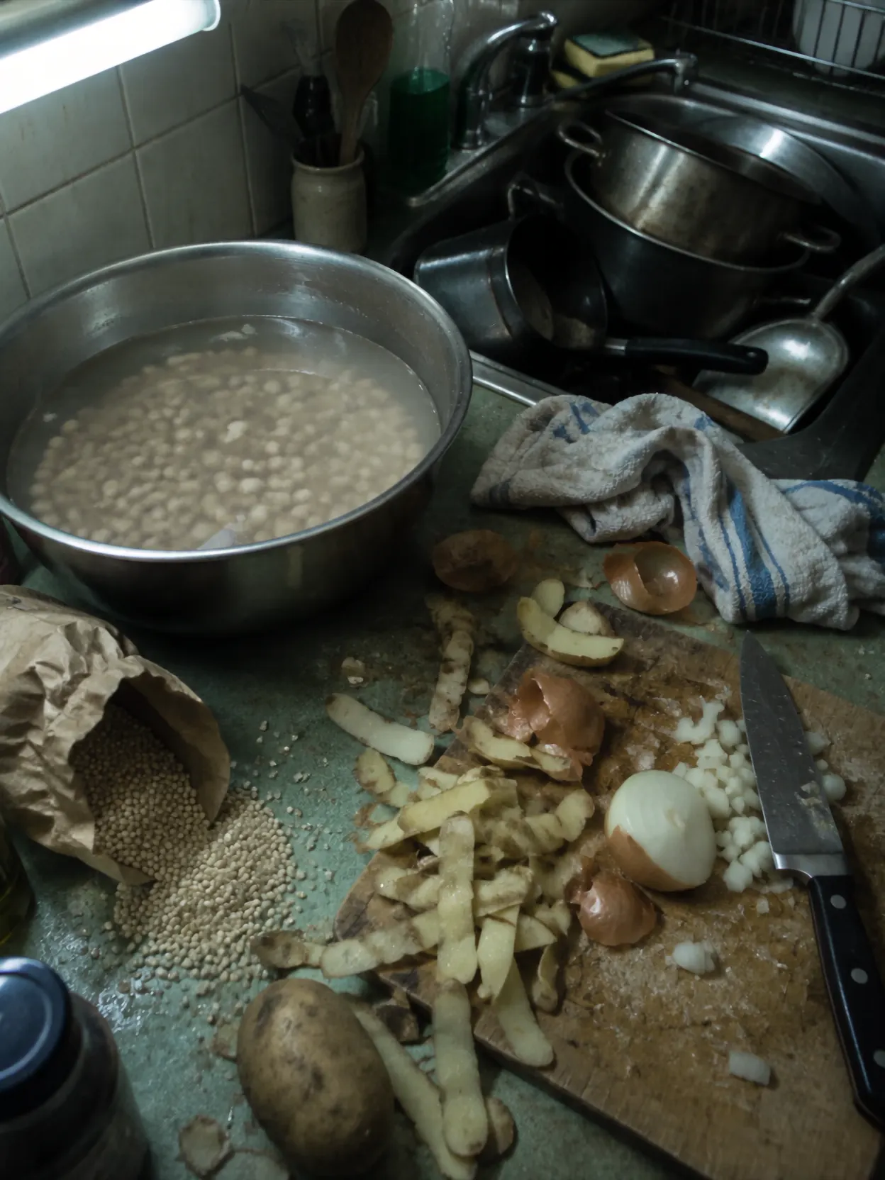 A cluttered kitchen counter late at night: beans soaking, barley spilling from a bag, potato peelings, onion skins and a knife.